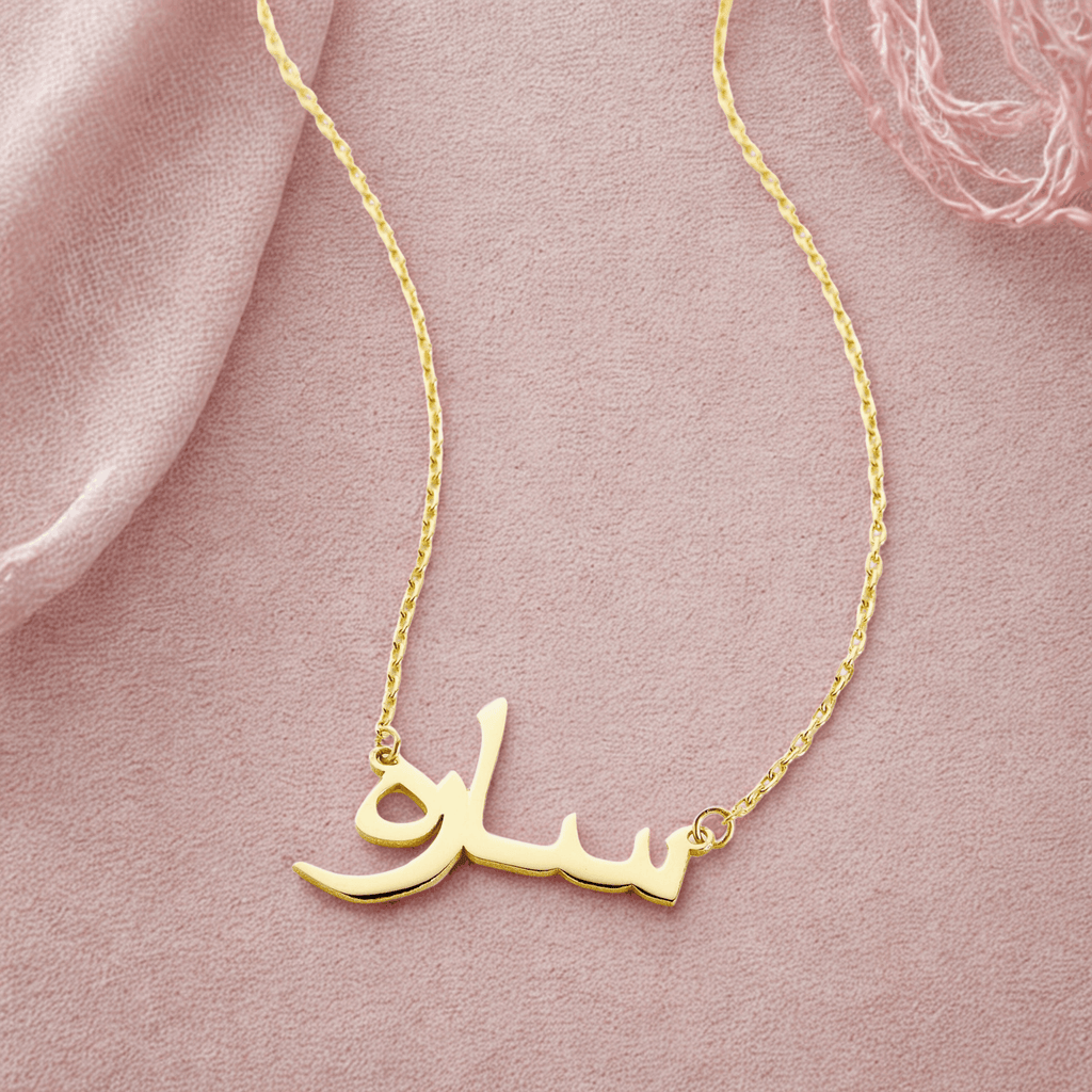 A delicate gold-plated chain necklace featuring a pendant in the shape of stylized Arabic script. The necklace is displayed against a pale blush background. The Arabic script appears to spell out a name, "Sarah".