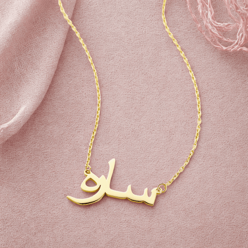 A delicate gold-plated chain necklace featuring a pendant in the shape of stylized Arabic script. The necklace is displayed against a pale blush background. The Arabic script appears to spell out a name, "Sarah".